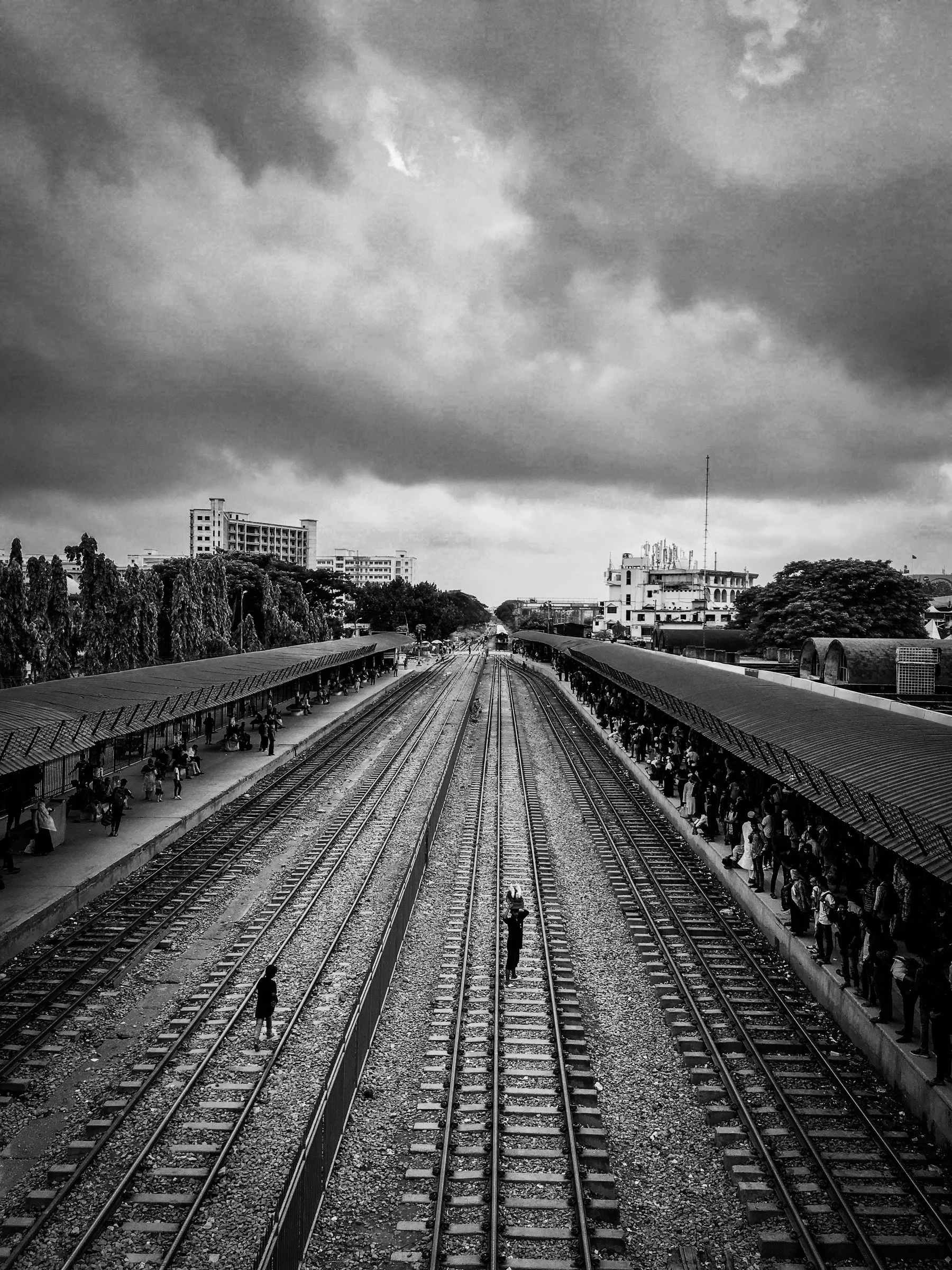 A figure carrying a load across empty railway lines, crowds waiting on both platforms
