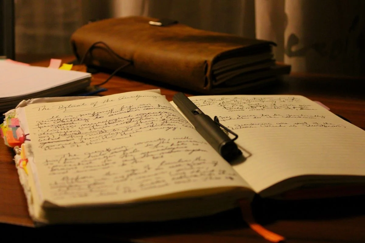 An open notebook filled with handwriting, a pen laid across the page, a leather bag behind it