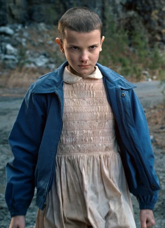 Eleven in a pink dress and blue jacket, standing on gravel